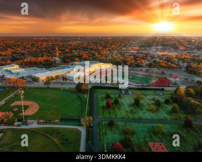 Blick aus der Vogelperspektive auf die Sportfelder der Naperville Central High School bei Sonnenuntergang, umgeben von farbenfrohen Herbstbäumen und glühender Skyline der Stadt. Stockfoto