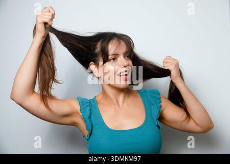Eine Frau zieht mit beiden Händen ihr langes Haar und lächelt in einem Innenbereich. Der Hintergrund ist schlicht und hell. Stockfoto