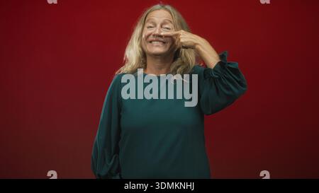Ältere Frau mit blonden Haaren zeigt den Finger an die Nase, lächelt, trägt grüne Bluse vor rotem Studio-Hintergrund; verspieltes Selbstvertrauen. Stockfoto
