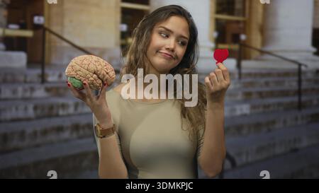Eine junge Frau im Freien, die ein Herz und ein Gehirn hält und die Wahl zwischen Liebe und Intellekt repräsentiert. Stockfoto