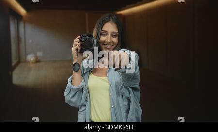 Junge hispanische Frau lächelt und hält die Kamera mit der Objektivkappe in der Hand, zeigt mit dem Finger auf das Objektiv im Studio; spielerische Verbindung. Stockfoto