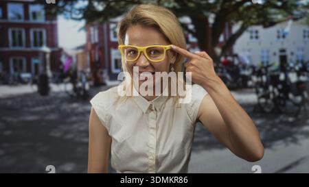 Die lächelnde junge blonde Frau gibt während des sonnigen Tages auf der belebten Stadtstraße eine Geste nach oben; Optimismus. Stockfoto