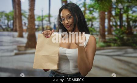Eine Frau, die eine Papiertüte hält, zeigt mit dem Finger auf die Kamera und macht auf der Straße eine Handbewegung mit dem Daumen auf sich selbst; selbstbewusste Einladung. Stockfoto