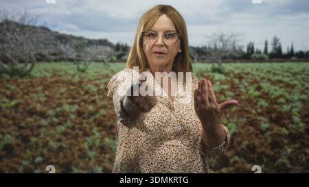 Frau zeigt mit gestreckter Hand und offener Handfläche die Hand in Richtung Kamera, trägt Brille und gemusterte Bluse im Wald; Entschlossenheit Warnung. Stockfoto