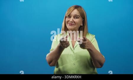 Die Frau zeigt beide Zeigefinger in einer Fingerpistolengeste, die Hände im Studio mit blauem Hintergrund zeigt; spielerisches Selbstvertrauen. Stockfoto