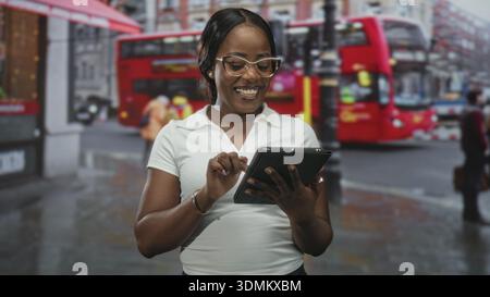 Frau, die mit den Fingern auf der regnerischen Stadtstraße in der Nähe des roten Doppeldeckerbusses auf das Tablet tippt; Freude am digitalen Fokus. Stockfoto
