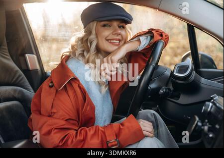 Sitzen und in die Kamera schauen, ausruhen. Glückliche blonde Frau sitzt in ihrem Elektroauto. Stockfoto
