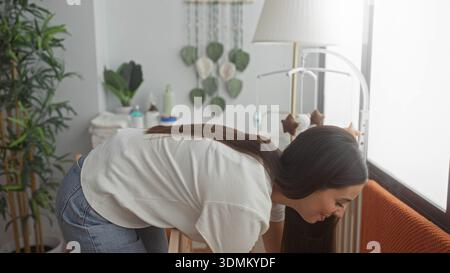 Frau, die das Baby im Schlafzimmer neben der Wiege und dem Mobilgerät liegend, sich über den Wickeltisch durch ein helles Fenster beugt; Zärtlichkeitsbindung. Stockfoto