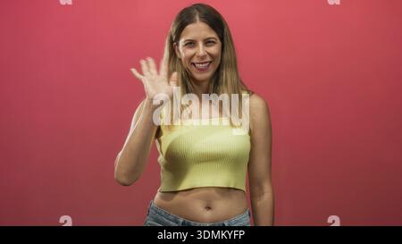 Frau winkte mit der Hand und zeigte Midriff in einem rosa Hintergrund und gelber Oberseite des Studios; freundliche Begrüßung. Stockfoto