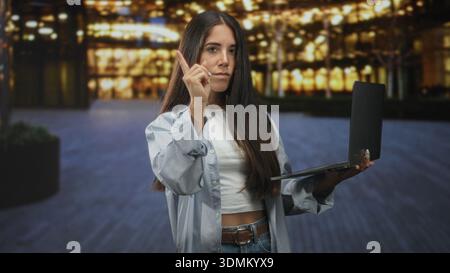 Frau, die den Laptop hält und den Finger nach vorne zeigt, zeigt bei Nacht vor dem beleuchteten gebäudeplatz einen Midriff; Fokusbestimmung. Stockfoto