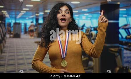 Arabische Frau mit Goldmedaille und Senfpullover zeigt den Finger in Richtung Kamera im Fitnessgebäude mit Laufbändern hinter sich, während sie lächelt; VI Stockfoto