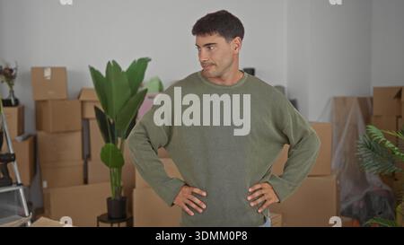Junger Mann steht mit Händen auf Hüften neben gestapelten Pappschachteln im Innenraum des Gebäudes und trägt einen olivfarbenen Pullover; Unsicherheit. Stockfoto