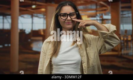 Frau Finger an Nase im Restaurant mit Brille und beigem Hemd, geschlossenen Augen und leichtem Lächeln; verspieltes Selbstvertrauen. Stockfoto