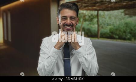 Der Mann im leeren Studio zeigt beide Zeigefinger auf seine Wangen, um ein verspieltes Grinsen zu erzeugen; Optimismus. Stockfoto