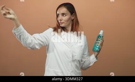Frau Ärztin und junge Rothaarige im weißen Labormantel hält eine blaue Flasche mit Kappe und zeigt den Zeigefinger in Richtung des Betrachters im Studio; Hygieneanleitung Stockfoto