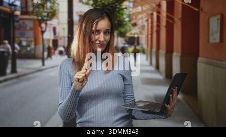 Frau, die einen Laptop hält, zeigt mit dem Zeigefinger auf eine Fußgängerzone, die von Bögen und Geschäften gesäumt ist, rote Haare und lässiger Pullover sichtbar; Selbstvertrauen. Stockfoto