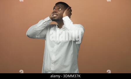 Mann mit Händen, die die Ohren im Studio bedecken, weißes Hemd, grimmend mit sichtbaren Ohrenschmerzen durch lautes Geräusch; Angstüberladung. Stockfoto