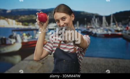 Frau, die roten Apfel mit Handschuhen am Hafenpier hält und Obst vor der Kamera präsentiert, während sie Schürze und gestreiftes Hemd trägt; gesunde Essfreude. Stockfoto