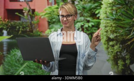 Blonde Frau mit Brille, die den Laptop hält, zeigt mit dem rechten Zeigefinger in den Innenhof mit grünen Pflanzen und Brunnen; gute Orientierungshilfe Stockfoto