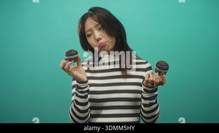 Frau hält zwei Schokoladenmuffins in beiden Händen, während sie in einem Studio Cupcakes vergleicht; zögerlich. Stockfoto