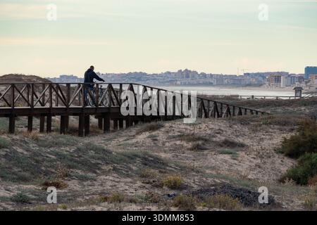 Mann mit seinem Fahrrad, der auf einem hölzernen Brückenweg durch die Sanddünen der Küste geht, die bei Sonnenuntergang mit goldener Stunde Licht und Dramat zum Strand des Ozeans führen Stockfoto
