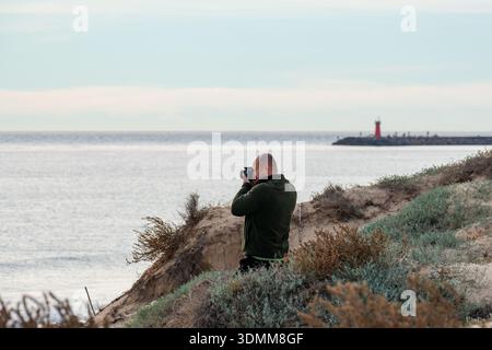 Fotograf, der ein Foto von einer Küstenlandschaft mit einem roten Leuchtturm auf einem Steg in der Ferne macht Stockfoto