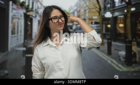 Die Frau zeigt mit dem Finger auf den Tempel auf einer Straße in der Stadt, trägt ein weißes Hemd und eine Brille; Ärgernis, zögerlich, Zweifel. Stockfoto