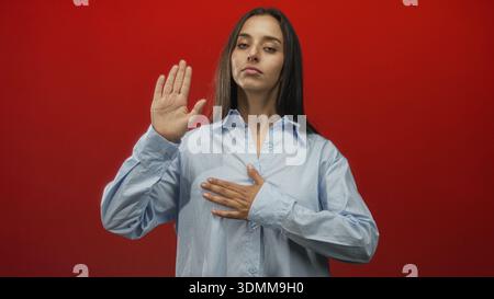 Frau hebt die Hand in die Kamera im roten Studio, während sie die andere Hand als Eid-Geste auf die Brust legt; feierliches Pfand. Stockfoto