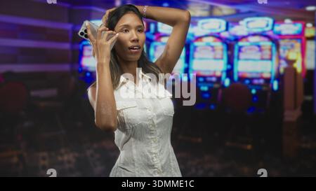 Frau, die das Smartphone am Ohr hält, redet und berührt Haare in einem Casino-Gebäude, ärmelloses weißes Kleid und Armband sichtbar; Spaß. Stockfoto
