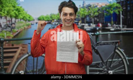 Mann im orangefarbenen Jumpsuit hält ein gefaltetes Papier mit beiden Händen an einer Straßenkanalbrücke, beißt auf die Lippe und hebt leicht die Faust; Trotz. Stockfoto