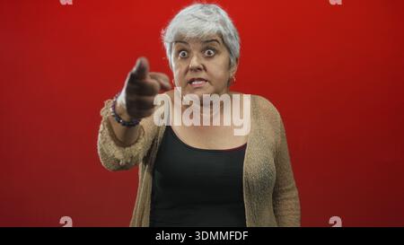 Frau zeigt mit gestreckter Hand und starkem Ausdruck mit dem Finger auf die Kamera, trägt Strickjacke und Tanktop vor rotem Studiohintergrund; beschimpft die Schuld. Stockfoto