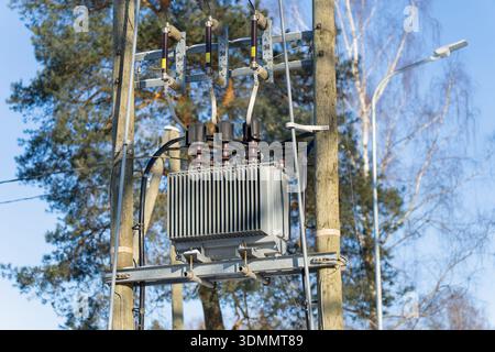 An Masten montierter elektrischer Transformator mit Isolatoren und Schaltanlagen an hölzernen Versorgungsmasten, Freileitungen, Straßenlaternen und Bäumen im Hintergrund Stockfoto