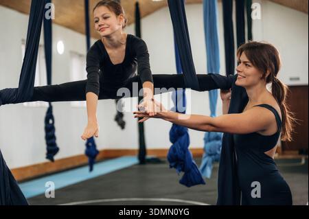 Zwei Personen, Sport. Eine Lehrerin hilft einem Kind, Luft-Yoga auf Hängematte-Gurten in einem Fitnessstudio zu üben. Stockfoto