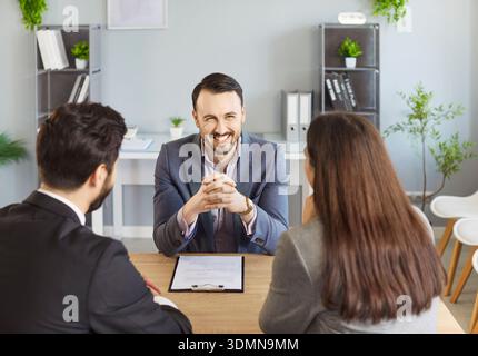 Lächelnder Geschäftsmann, der am Schreibtisch sitzt und mit zwei Kunden während der Beratung im modernen Büro spricht. Stockfoto