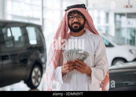 Ein arabischer Geschäftsmann hält Bargeld in einem modernen Autohaus. Stockfoto