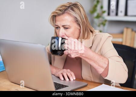 Geschäftsfrau, Die Bei Laptop Im Büro Arbeitet Und Während Workday Kaffee Aus Der Schwarzen Tasse Trinkt Stockfoto