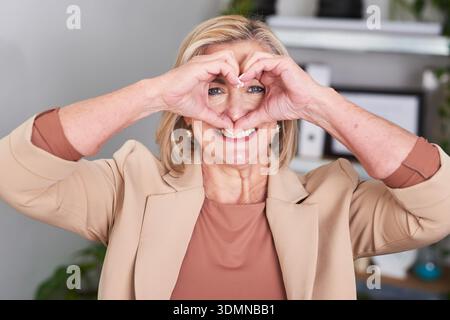 Lächelnde Seniorin Macht Herzförmige Hände Bei Der Arbeit, Drückt Heute Liebe, Freundschaft Und Positivität Aus Stockfoto