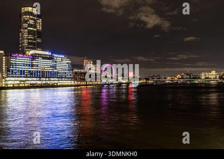 Nächtlicher Blick auf die Londoner Stadtlandschaft von der anderen Seite der Themse aus mit beleuchteten Gebäuden und dem leuchtenden London Eye unter einem dunklen, bewölkten Himmel. Stockfoto