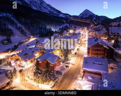 Blick aus der Vogelperspektive auf das bezaubernde Dorf, eingebettet zwischen schneebedeckten Dächern, beleuchteten Straßen und majestätischen Berggipfeln unter einem Dämmerungshimmel, Claviere, Stockfoto