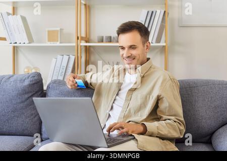 Lächelnder Mann, der online mit Laptop und Kreditkarte auf einem komfortablen Sofa einkaufen wollte Stockfoto
