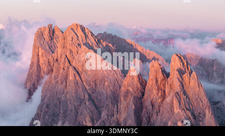 Aus der Vogelperspektive der zerklüfteten Gipfel, die von der rosafarbenen Morgendämmerung geküsst werden und durch ein Meer aus wirbelnden Wolken durchdringen, Seceda, Italien. Stockfoto