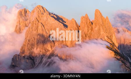 Aus der Vogelperspektive auf die zerklüfteten Gipfel von Seceda, die durch ein Meer aus weichen, ätherischen Wolken durchdringen, beleuchtet vom warmen Schein der untergehenden Sonne, Dolomiten, Ita Stockfoto