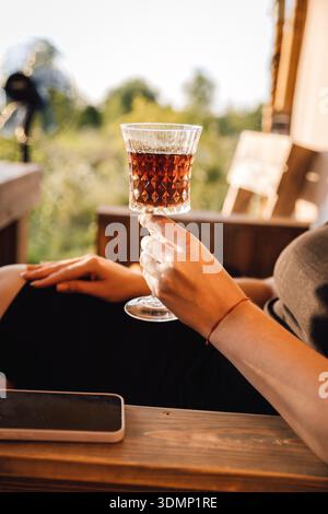 Hand in der Hand mit Kristall Cocktailglas auf sonniger Terrasse mit Smartphone Stockfoto