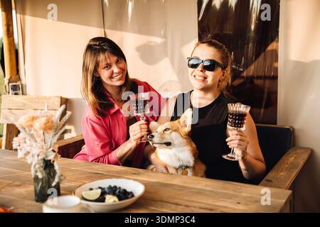 Zwei Frauen toasten Wein mit Corgi auf der sonnigen Außenterrasse Stockfoto