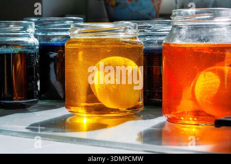 Nahaufnahme eines ostereiers in einer gelben und orangen Lebensmittelfarbstofflösung in einem Glasgefäß auf einem sonnendurchfluteten Küchentisch Stockfoto