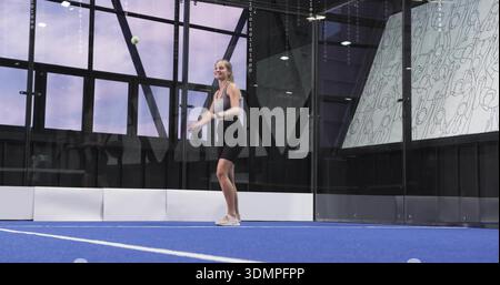 Servierfrau in schwarzem sportlichem Outfit auf blauem Platz, Glaswänden und kleinem Ball in der Luft Stockfoto
