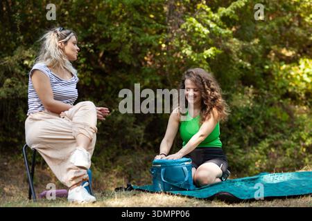 Frauen teilen sich beim Camping im Freien einen festen Moment. Der eine sitzt auf einem Stuhl und führt Gespräche, während der andere auf seinem Campingplatz Essen zubereitet Stockfoto