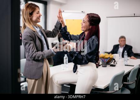 Zwei Geschäftsfrauen arbeiten fröhlich während eines produktiven Meetings in einem modernen Büroumfeld mit anderen Fachleuten im Hintergrund. Stockfoto