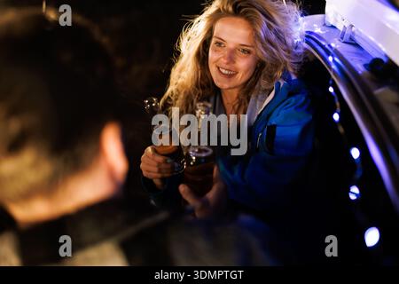 Freunde genießen bei einem gemütlichen Treffen im Freien Getränke unter dem Nachthimmel, während sie in einem von sanften Lichtern beleuchteten Auto sitzen Stockfoto