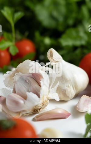 Knoblauchzehen und eine ganze Knolle sitzen neben Kirschtomaten auf einem Schneidebrett, umgeben von frischen Kräutern. Stockfoto